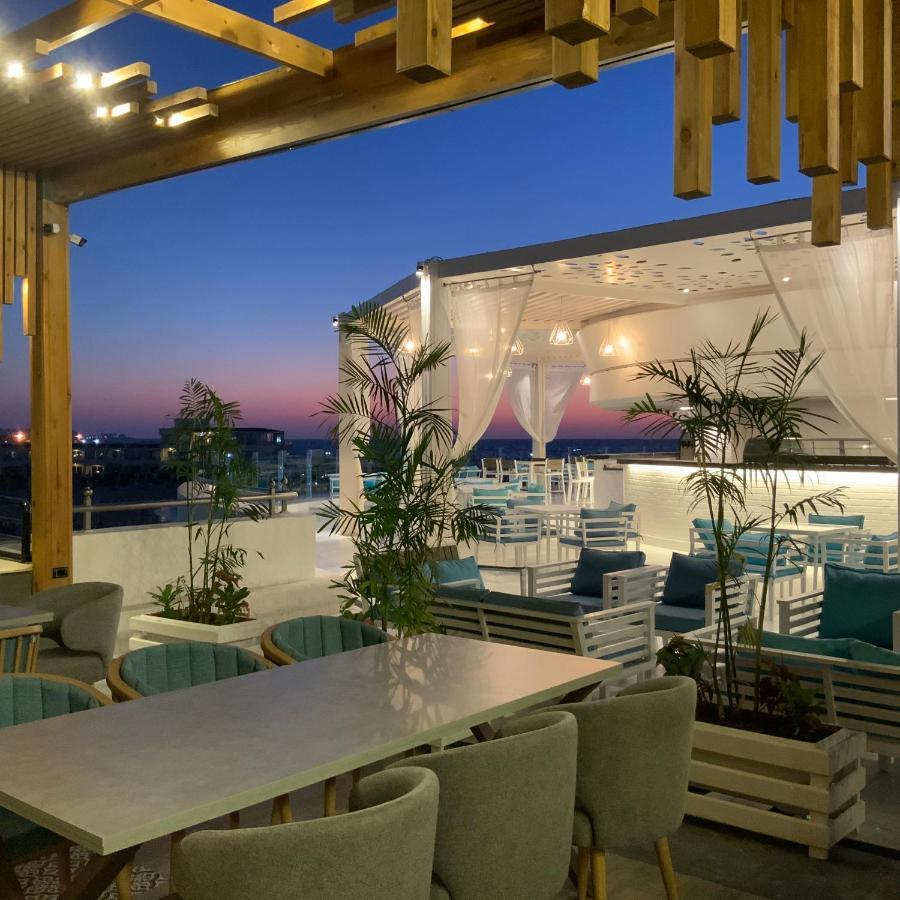 Sunrise Alex Avenue Hotel Alexandria Exterior foto The photo shows a stylish outdoor dining area or terrace set against a beautiful sunset. There are several tables and chairs arranged neatly under a covered structure, with a backdrop of soft white curtains. The space is decorated with potted plants,