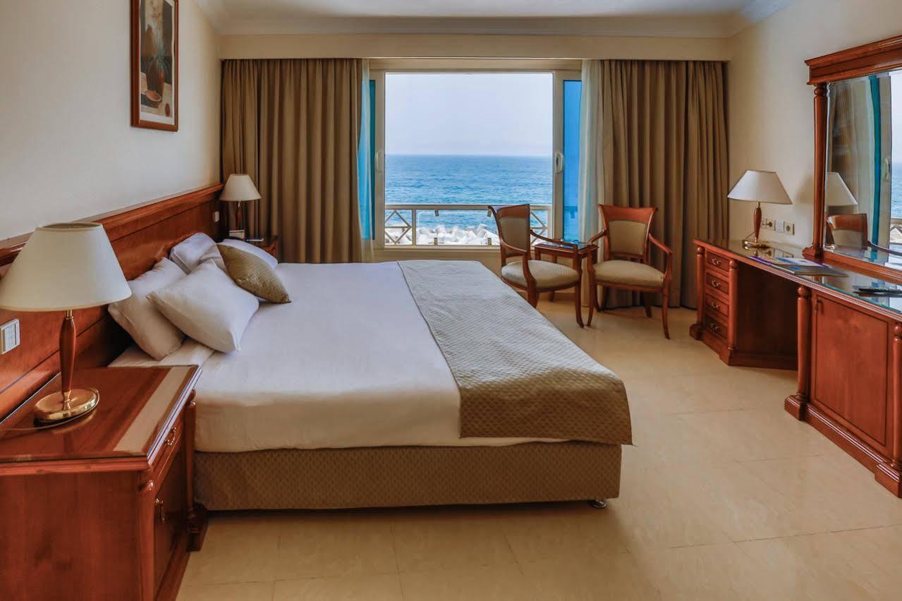 Sunrise Alex Avenue Hotel Alexandria Exterior foto The photo shows a hotel room featuring a large bed with white linens and decorative pillows. There are two wooden chairs positioned near a window, which offers a view of the ocean. The room has beige walls and light-colored curtains that frame the wi