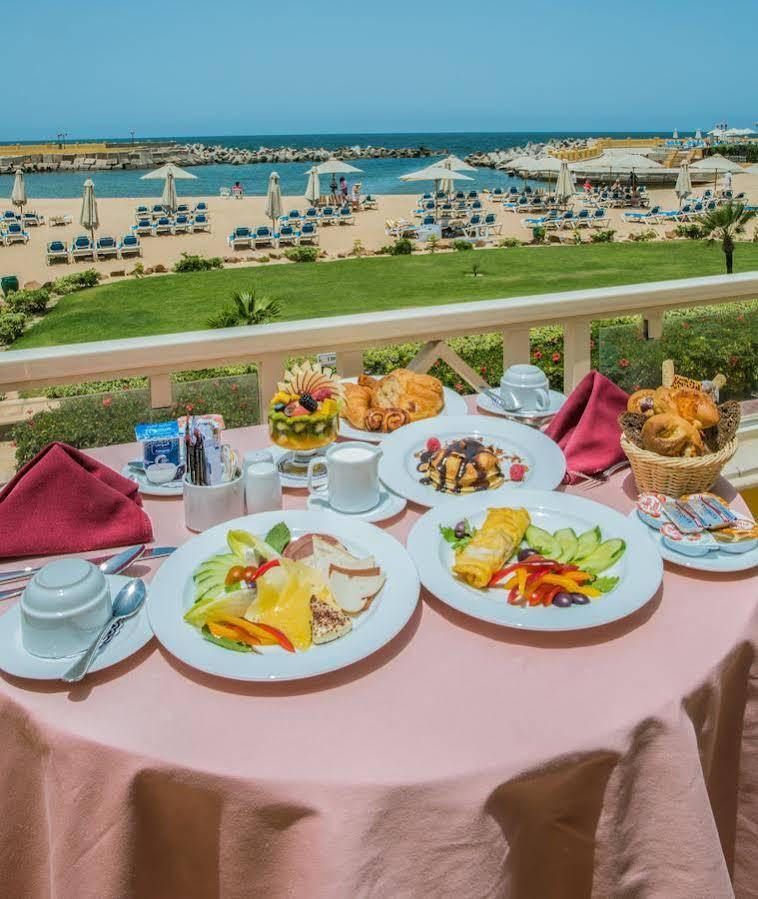 Sunrise Alex Avenue Hotel Alexandria Exterior foto The photo shows a beautifully set breakfast table on a balcony with a scenic view of a beach. There are several plates of food arranged attractively. One plate features an assortment of fruits, including slices of melon, berries, and other colorful f