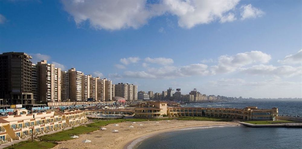 Sunrise Alex Avenue Hotel Alexandria Exterior foto The photo shows a scenic coastal view featuring a sandy beach and a curved shoreline. In the foreground, there are several sun loungers and a landscaped area. Beyond the beach, there are multiple high-rise buildings lining the waterfront, indicating 