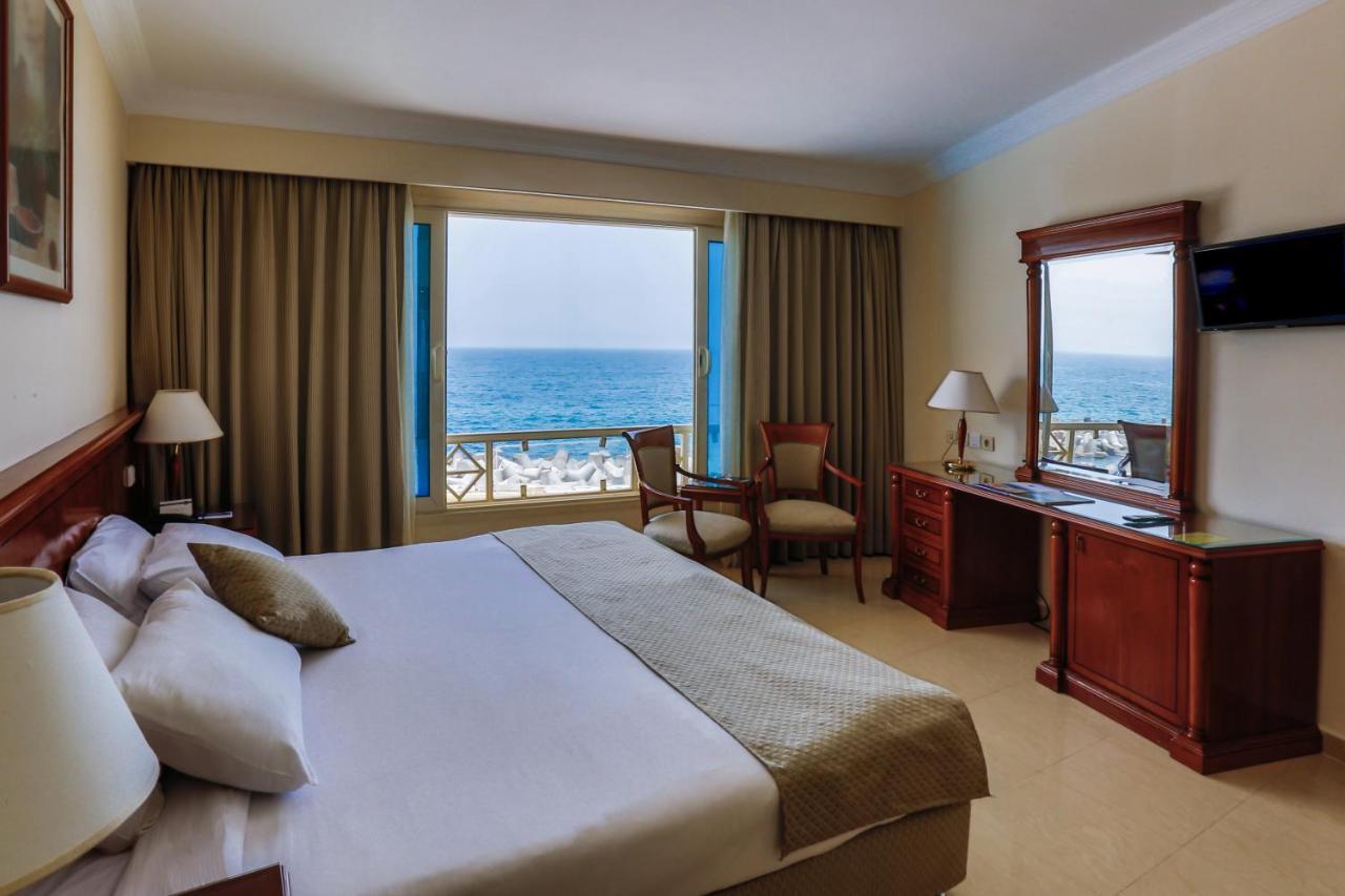 Sunrise Alex Avenue Hotel Alexandria Exterior foto The photo shows a hotel room with an ocean view. The room features a large bed with neutral-colored bedding and pillows. There are curtains covering the windows, allowing plenty of light to enter. Beside the bed, there is a wooden desk and a chair po