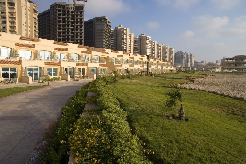 Sunrise Alex Avenue Hotel Alexandria Exterior foto The photo depicts a scenic view of a shoreline area with modern buildings in the background. In the foreground, there is a well-maintained green lawn, bordered by a pathway. The buildings appear to be multi-story residential or commercial structures,