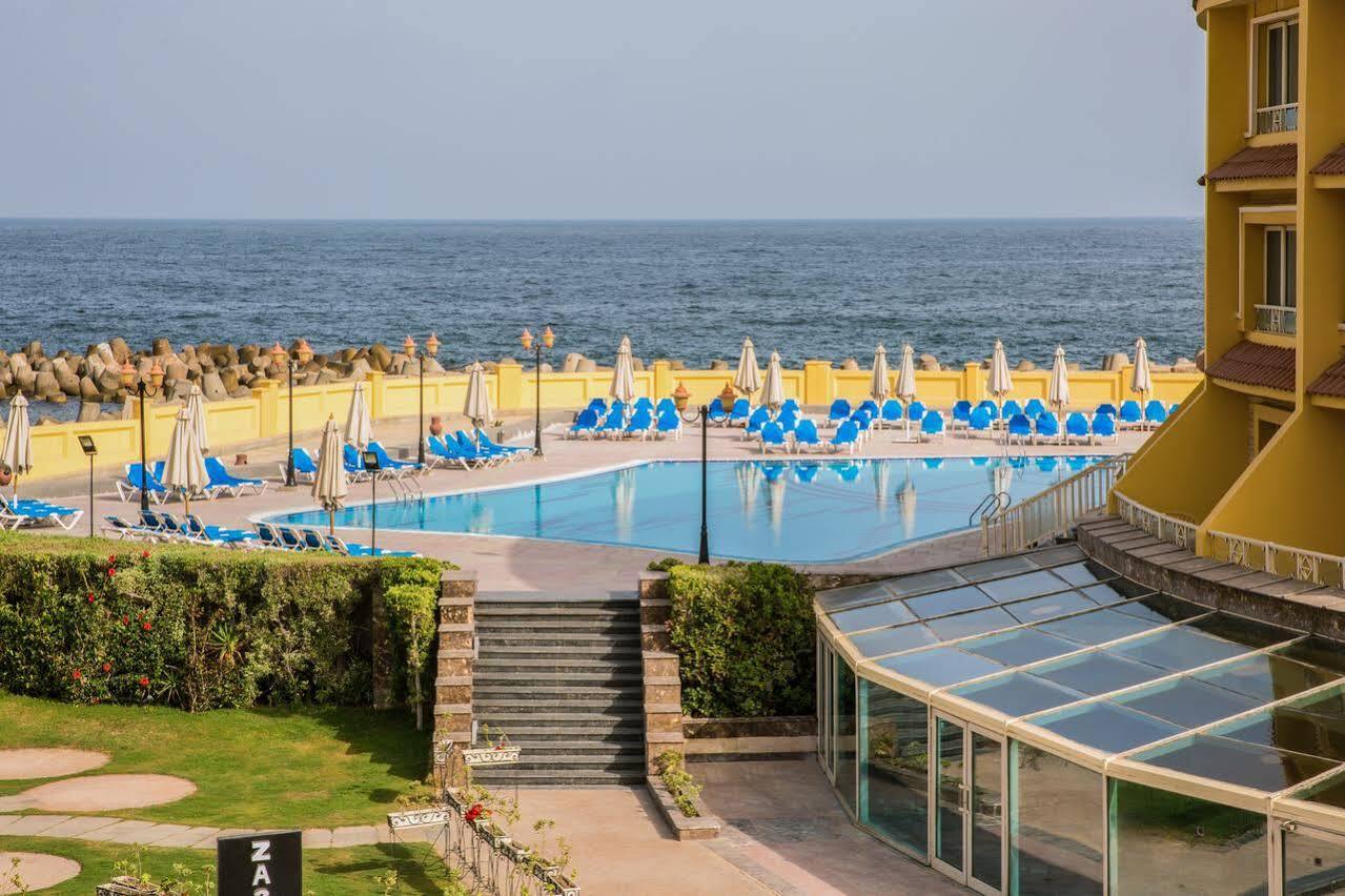 Sunrise Alex Avenue Hotel Alexandria Exterior foto The photo depicts a scenic view of a pool area adjacent to the ocean. In the foreground, there is a large swimming pool surrounded by numerous blue lounge chairs and umbrellas, indicating a relaxing setting. There are steps leading down from a grassy