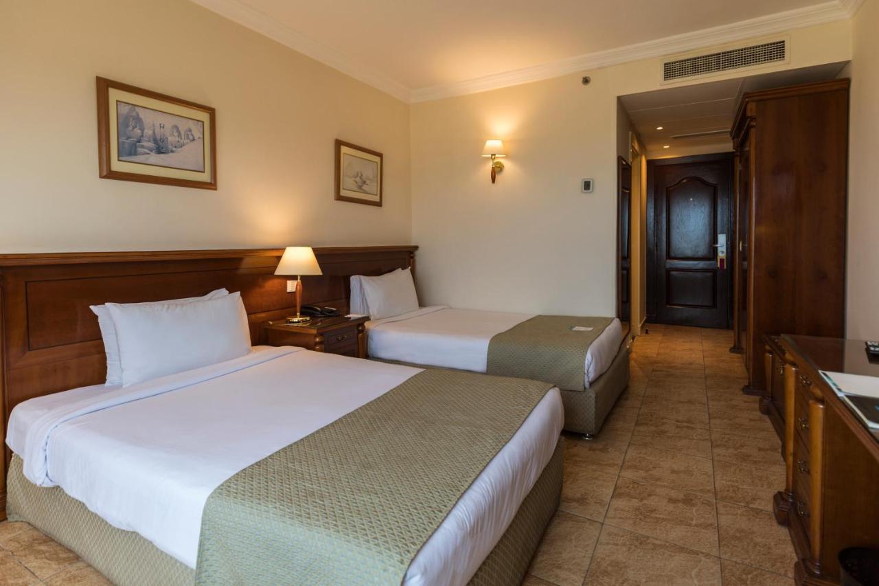 Sunrise Alex Avenue Hotel Alexandria Zimmer foto The photo shows a hotel room featuring two twin beds with light-colored bed linens and green decorative blankets. There's a bedside table with a lamp next to one of the beds. The walls are painted a light color, and there are two framed pictures hang