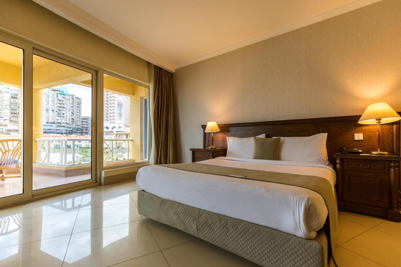 Sunrise Alex Avenue Hotel Alexandria Zimmer foto The photo shows a spacious hotel room featuring a large bed with white linens and decorative pillows. There are wooden nightstands with lamps on both sides of the bed. The walls are painted in neutral tones, and the floor is tiled. Large windows prov